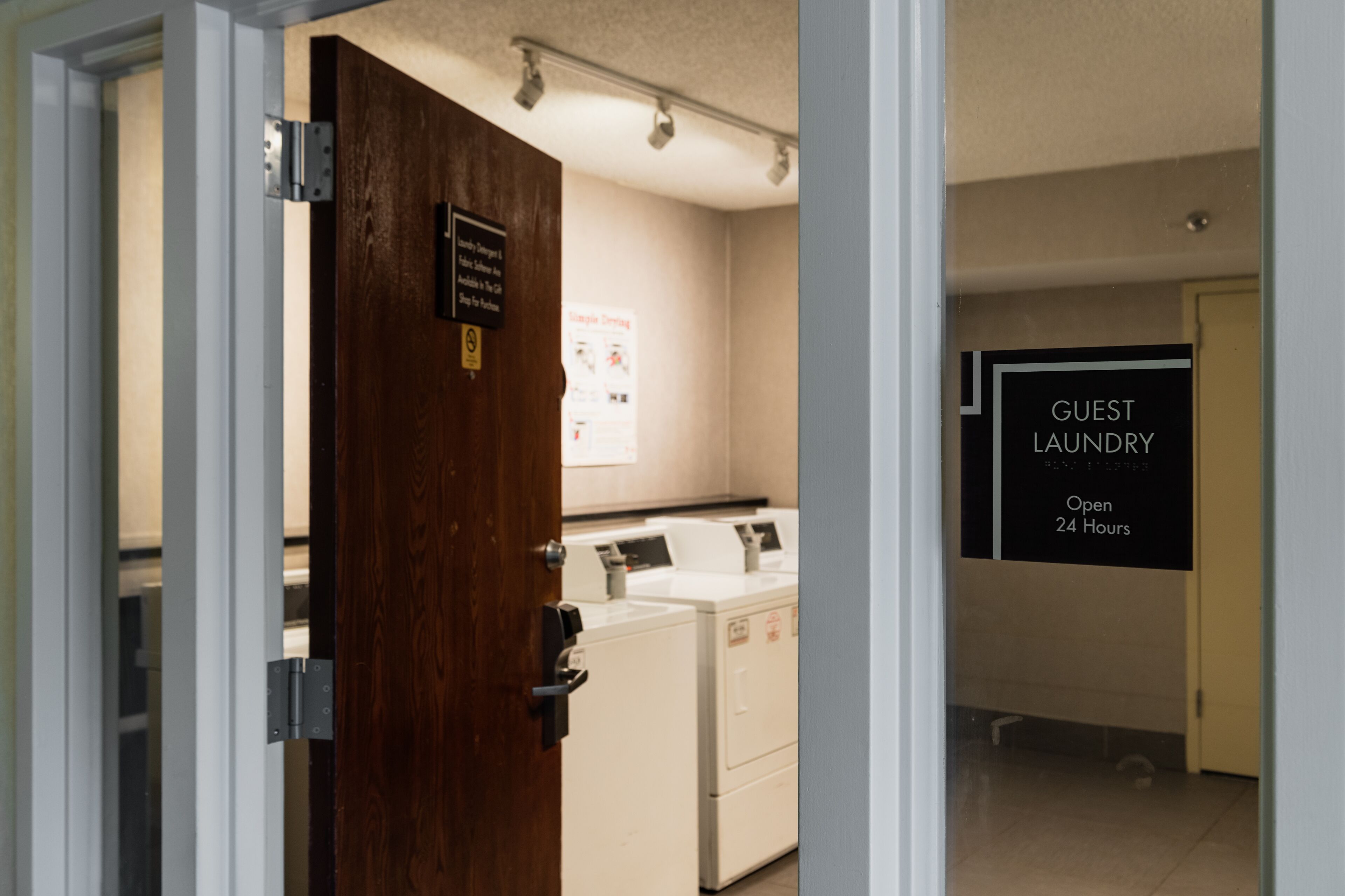 A hotel room with a wooden door, a sign indicating guest laundry facilities open 24 hours, and a glimpse of a laundry area with washing machines.