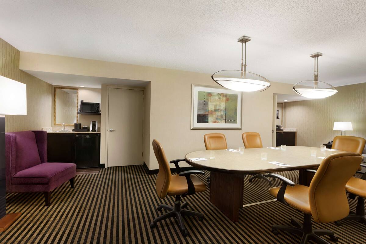 A hotel room with a conference table, chairs, a purple armchair, a kitchen area, and artwork on the wall.