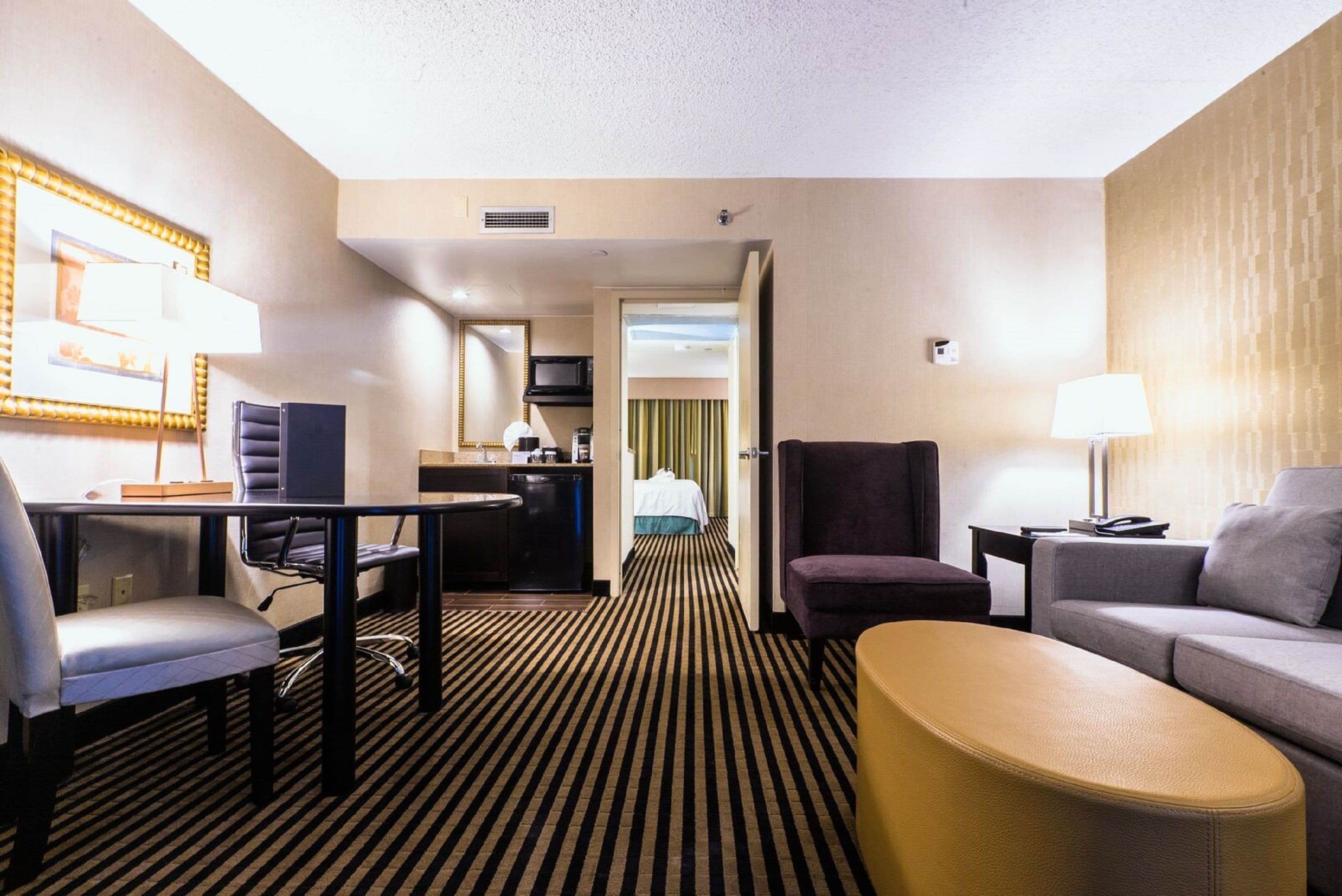 Habitación de hotel con alfombra a rayas, un sofá, una silla, una mesita, un escritorio con silla, un televisor, una lámpara y una cama al fondo.