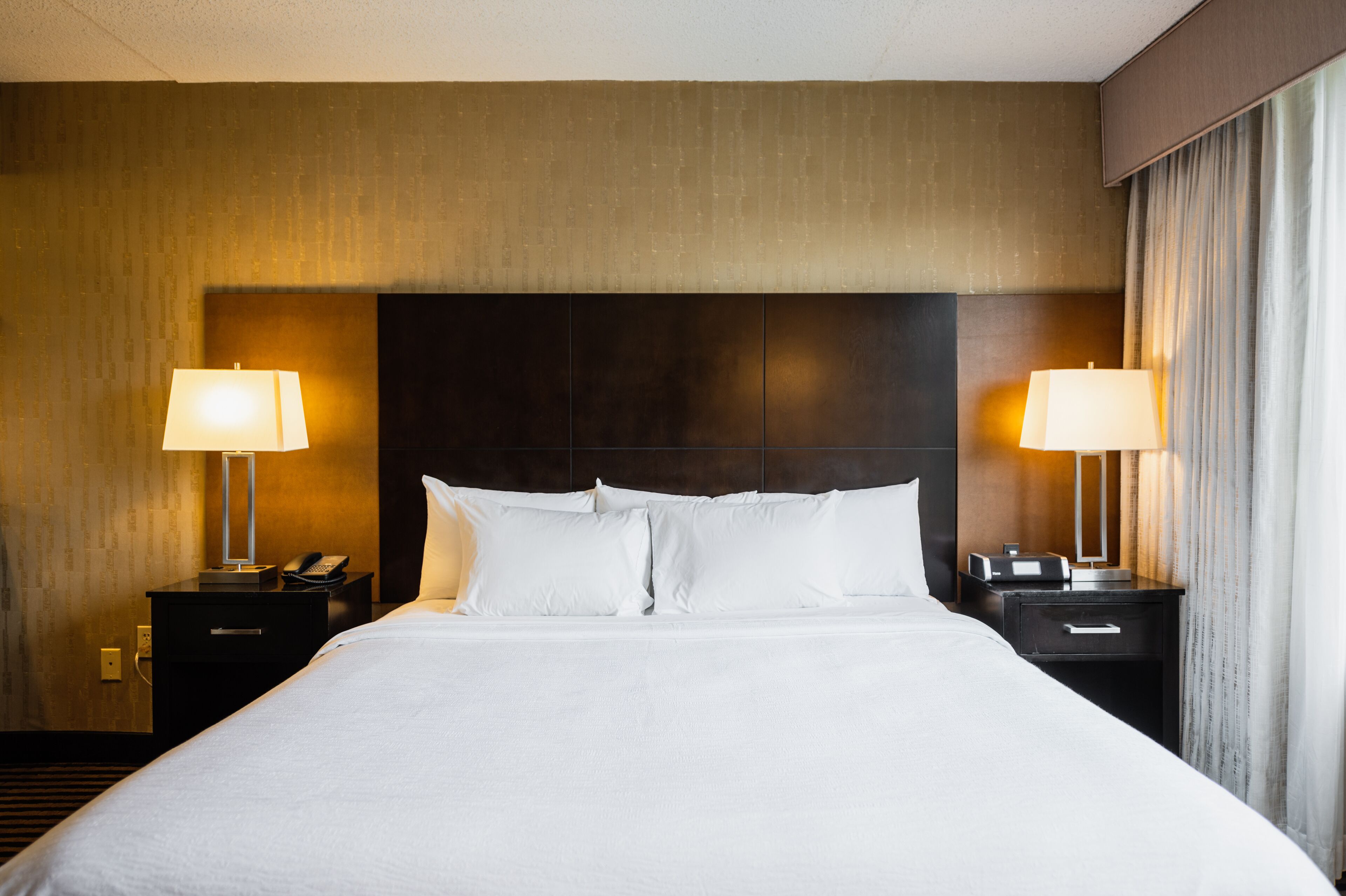 A hotel room with a large bed, two bedside tables with lamps, a dark headboard, and a curtain on the window.