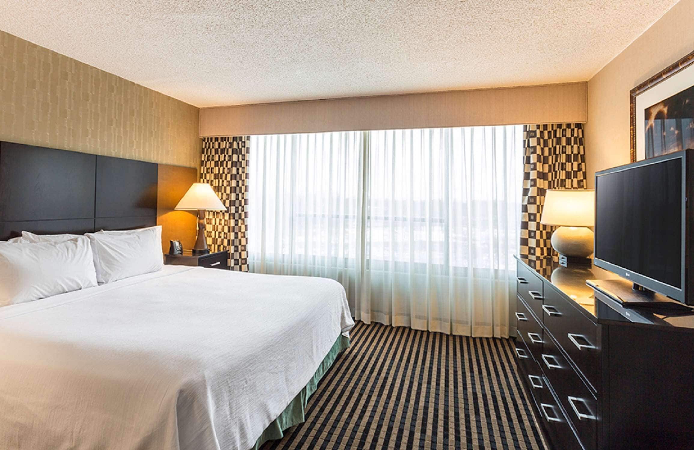 A hotel room with a large bed, a television, a desk with a lamp, and a view of the city through large windows.
