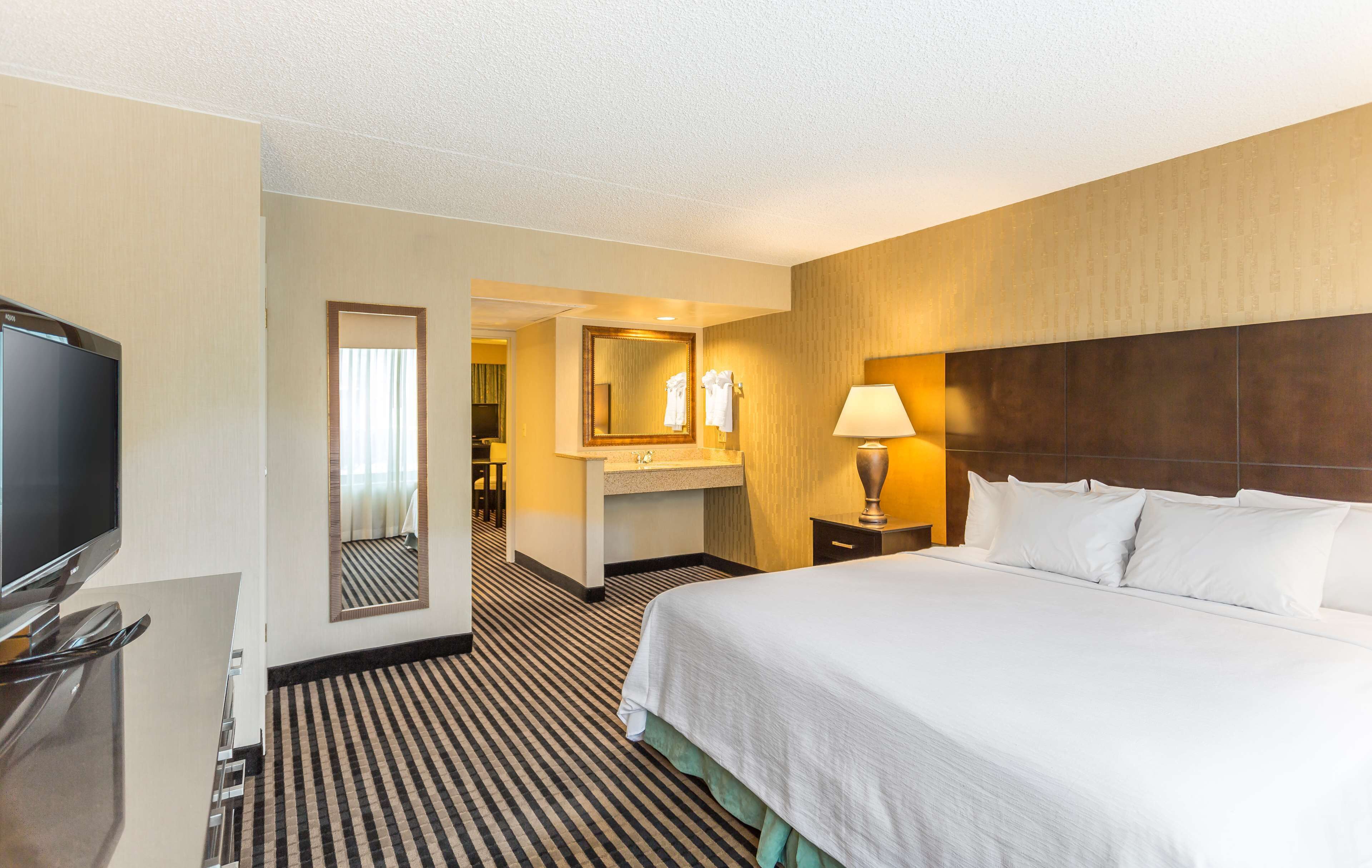 A hotel room with a large bed, a TV, a desk, a mirror, and a bathroom visible through an open door.