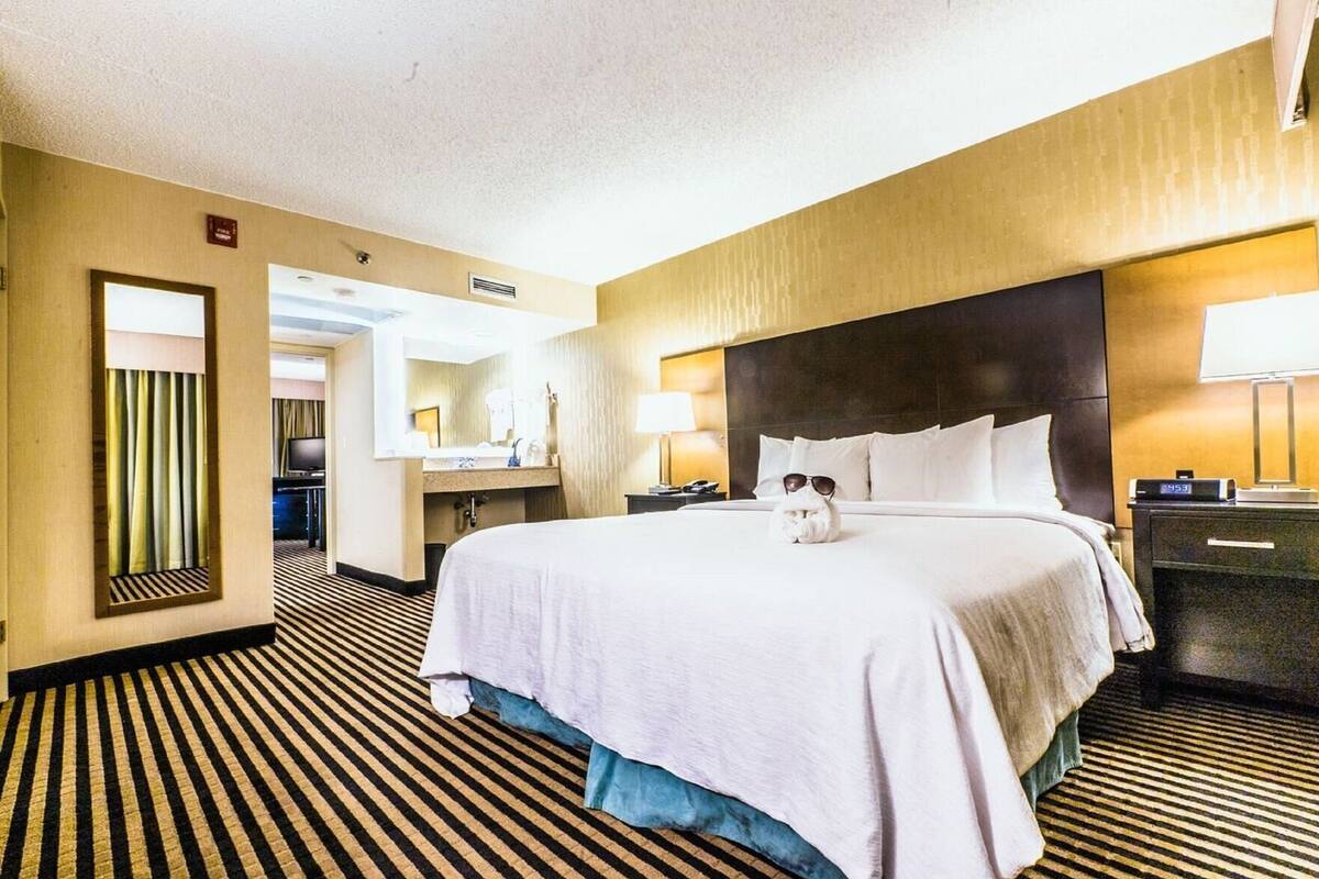 A hotel room with a large bed, a nightstand, a desk with a lamp, a mirror, and a bathroom visible through an open door.