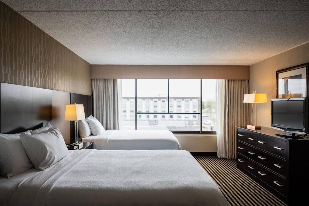 A hotel room with two beds, a TV, a dresser, and a large window with a city view.