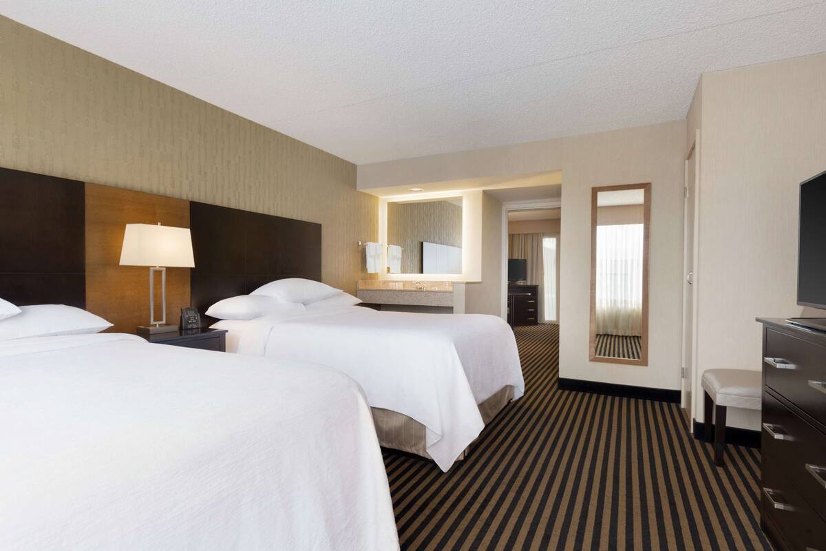 A hotel room with two beds, a desk, a dresser, a mirror, and a bathroom visible through an open door.