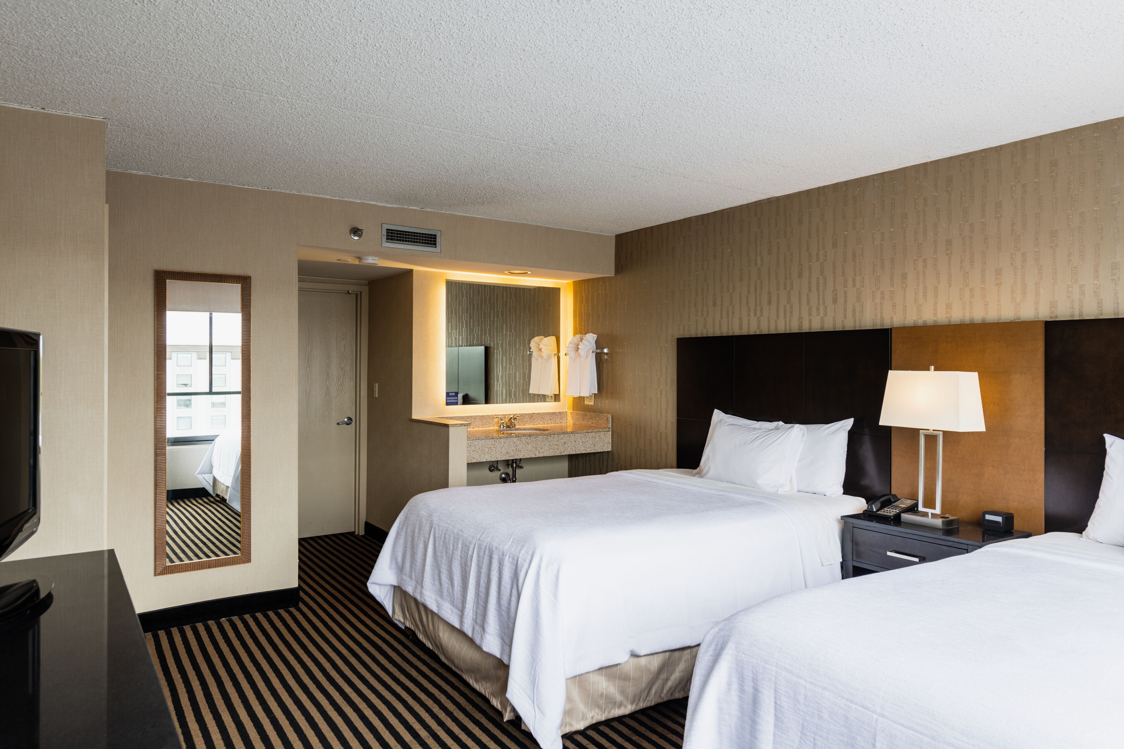 Une chambre d’hôtel avec deux lits, un grand miroir, une coiffeuse avec un lavabo, une télévision et une table de chevet avec une lampe.