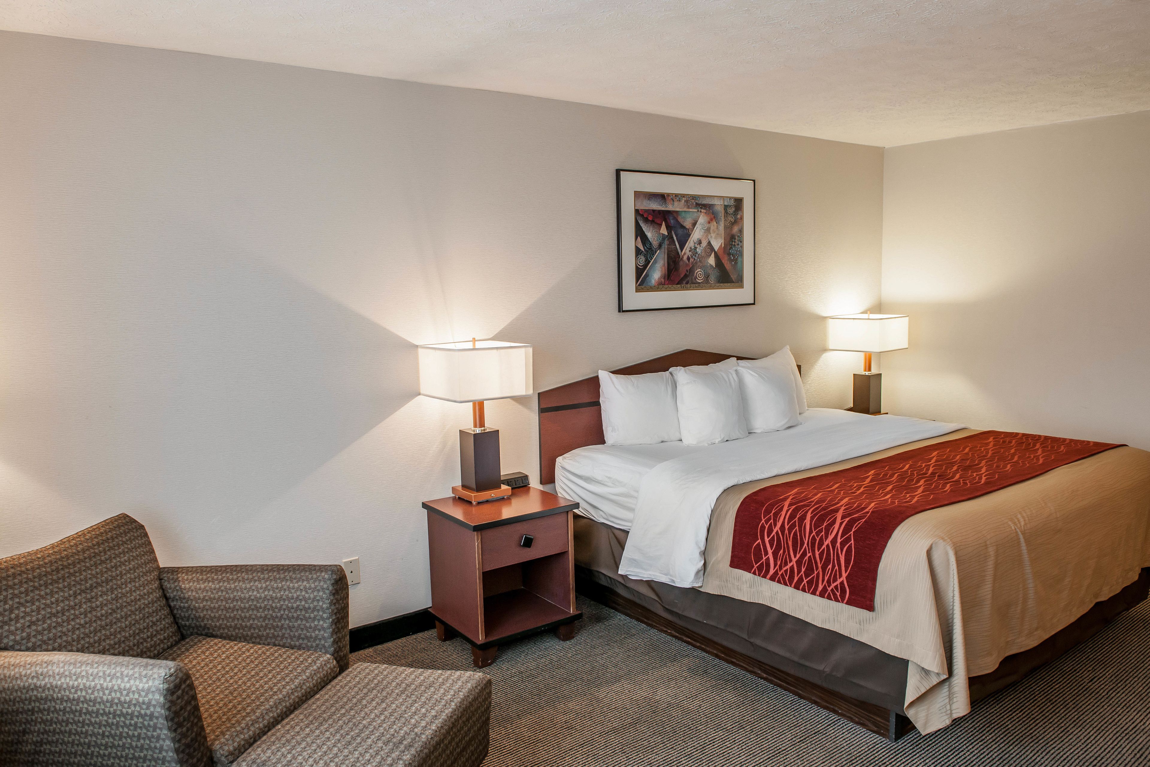 A hotel room with a bed, two bedside lamps, a small nightstand, a chair, and a painting on the wall.