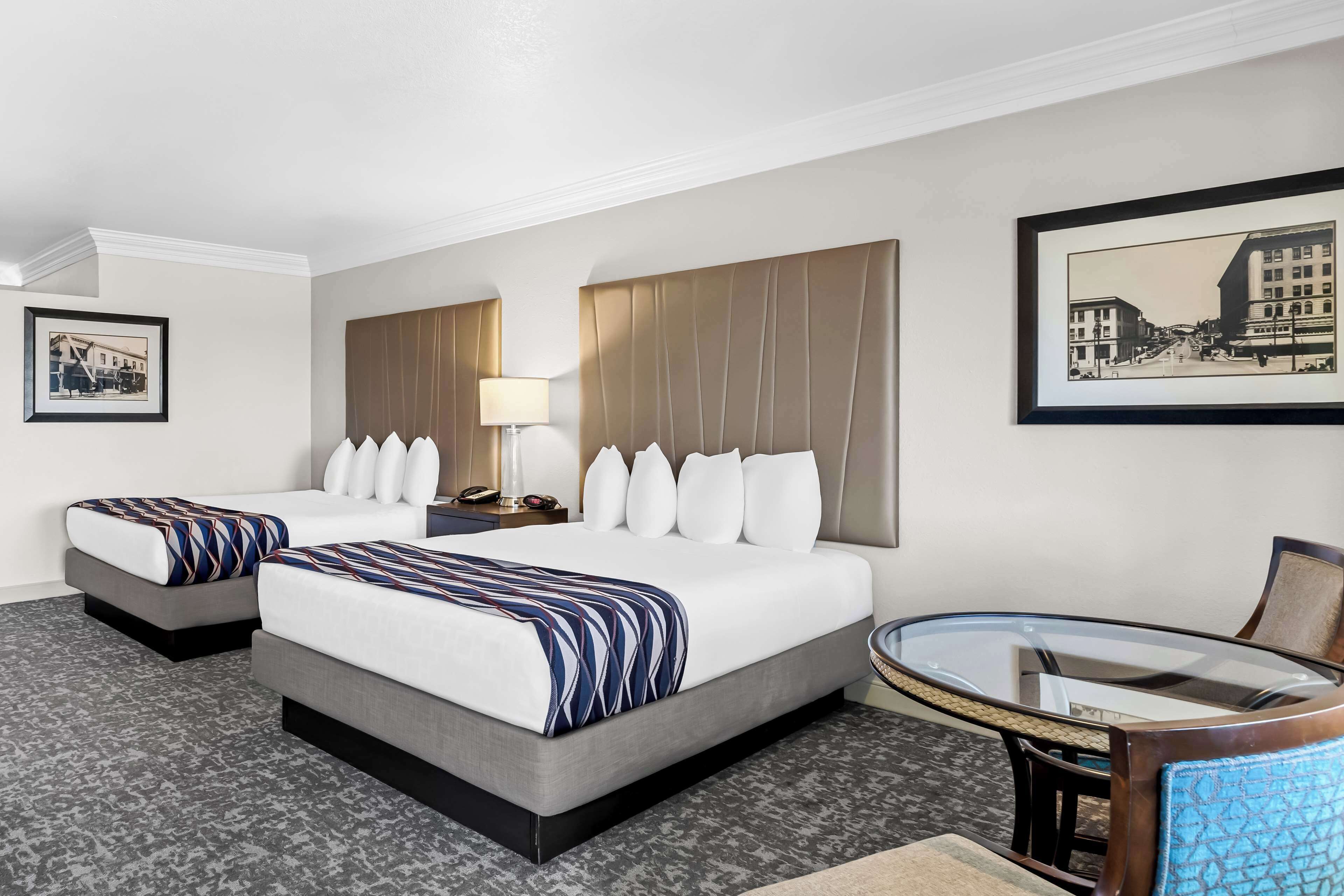 A hotel room with two beds, a round glass table, a chair, a nightstand with a lamp, and framed pictures on the walls.