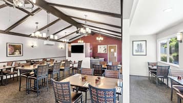 A spacious dining area with high ceilings, exposed beams, and modern chandeliers. Tables are arranged with chairs, and there are framed pictures on the walls.