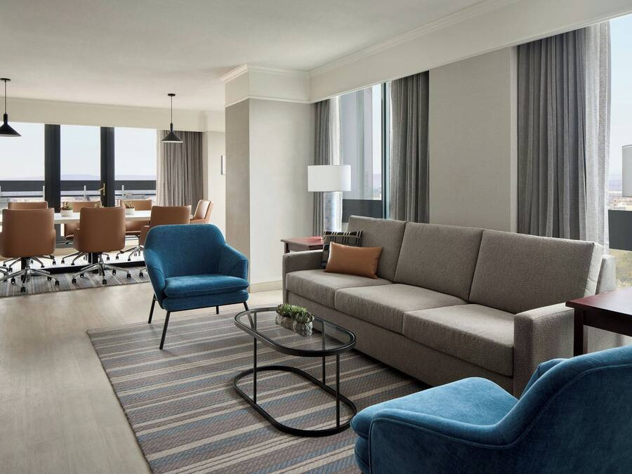 A modern hotel room with a sofa, two armchairs, a coffee table, and a dining area with chairs and a table.