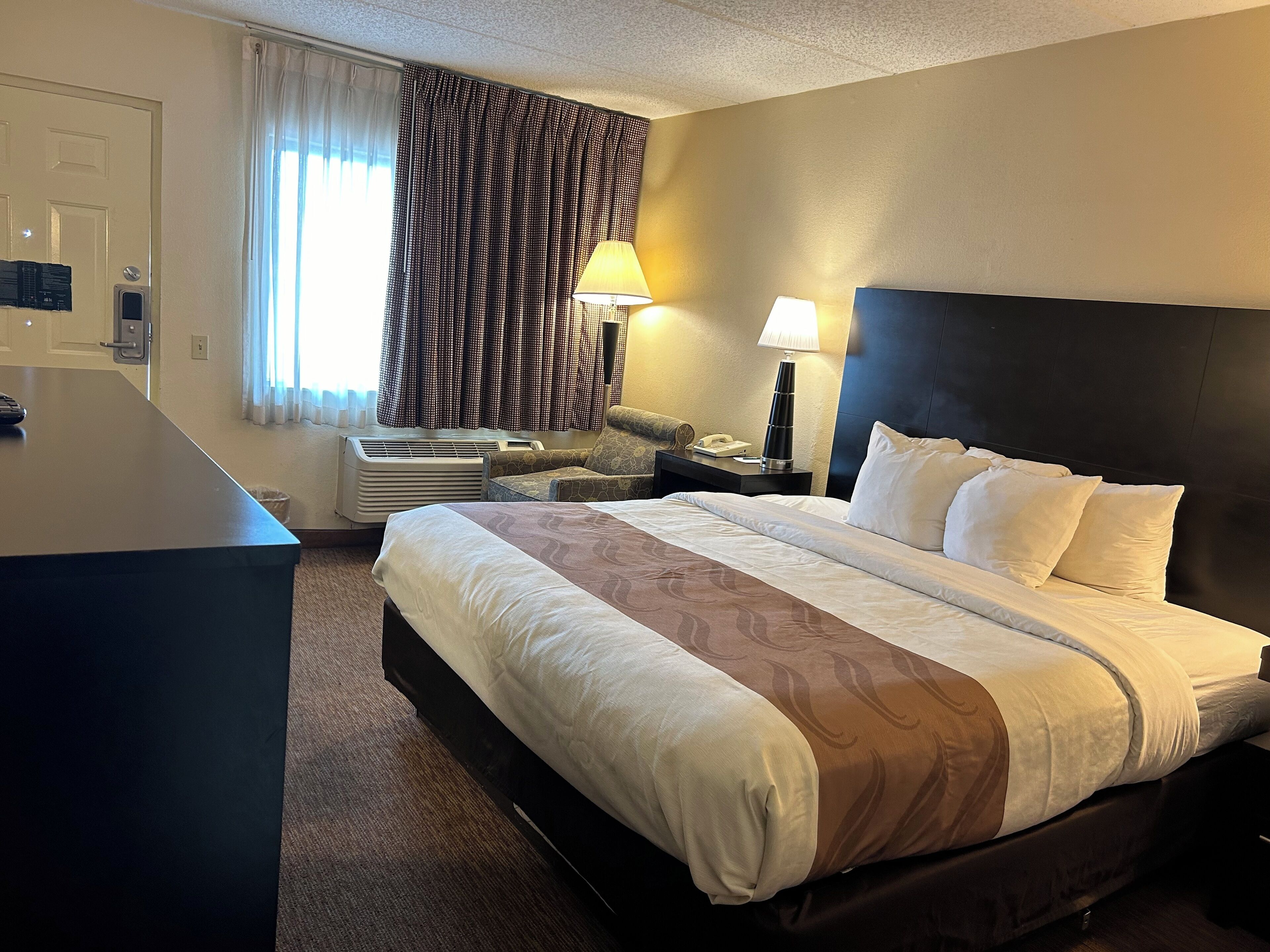 Une chambre d’hôtel avec un lit, un bureau, une chaise, une lampe et une fenêtre avec des rideaux.