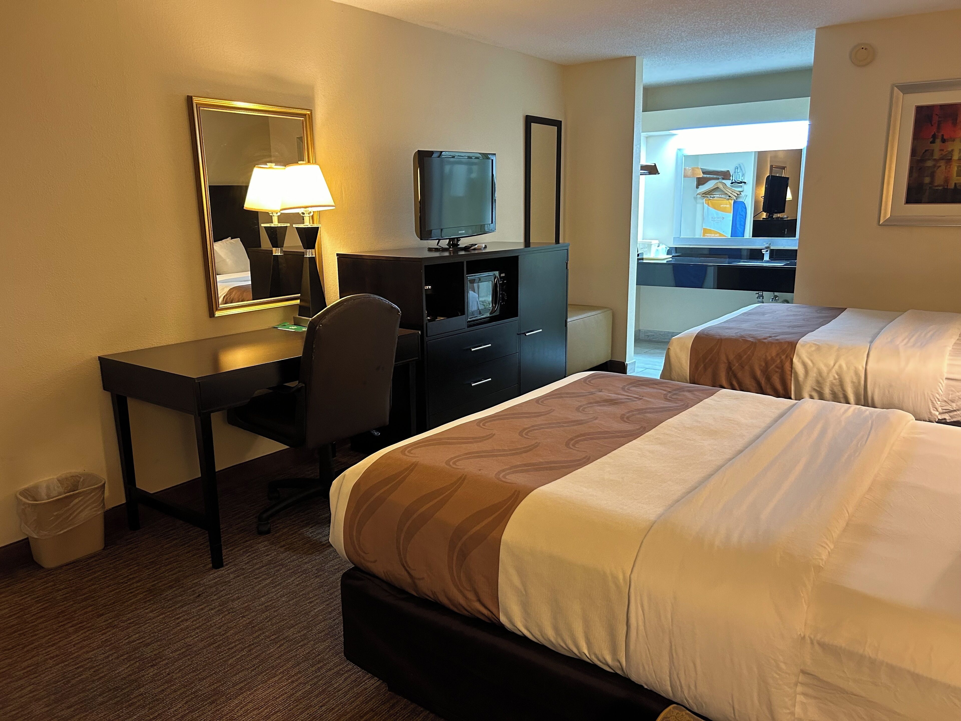 A hotel room with a desk, two beds, a television, and a mirror.