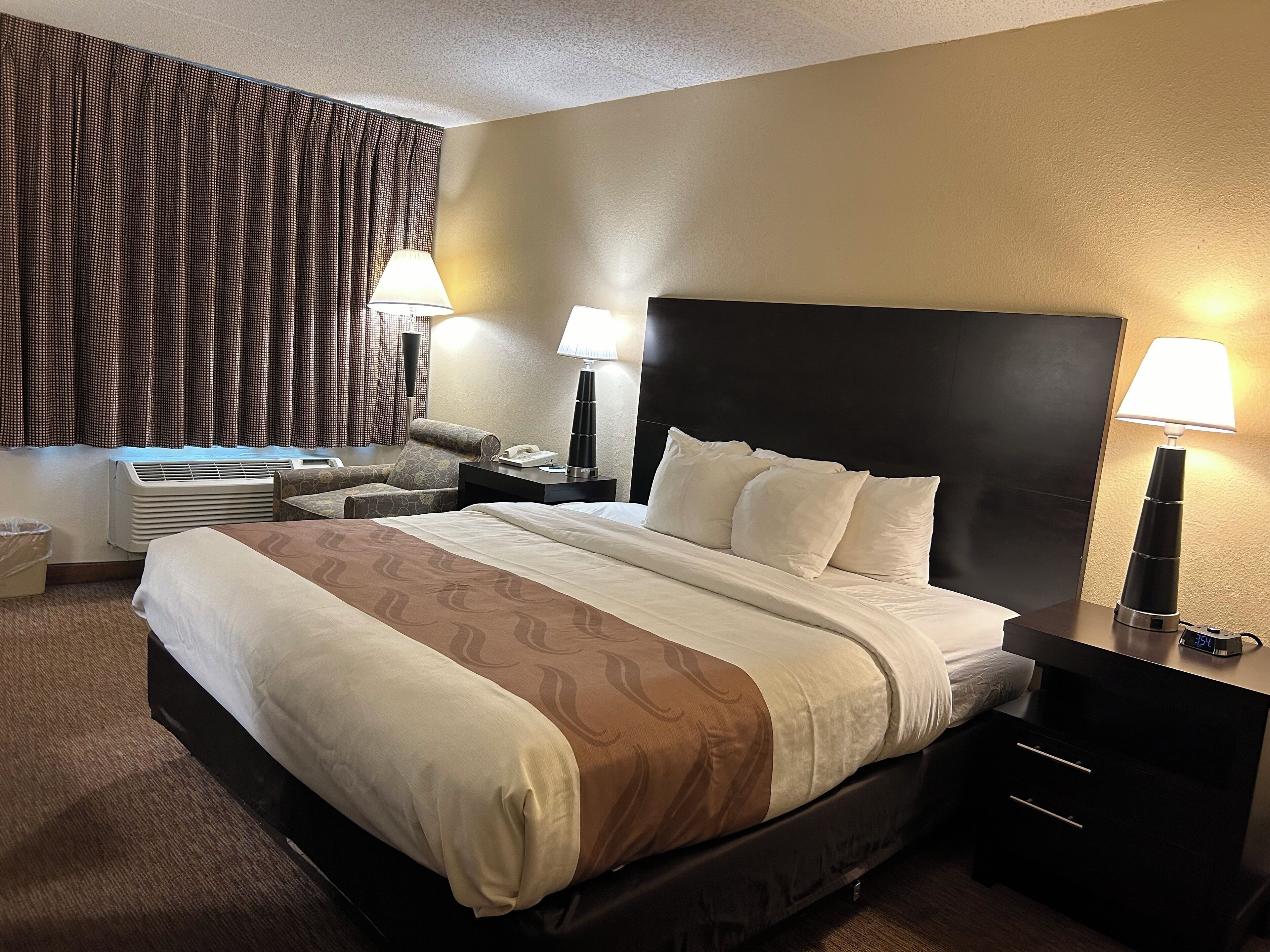 Una habitación de hotel con una cama grande, dos mesitas de noche con lámparas, una silla, un escritorio con teléfono y una ventana con cortinas.