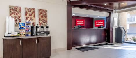 A hotel reception area with a coffee station, a red sign indicating free Wi-Fi in all rooms, and a view of the outside balcony.