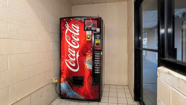 A Coca-Cola vending machine in a corner with a tiled floor and a window with a black frame. - Red Roof Inn Bordentown - McGuire AFB (Bordentown)
