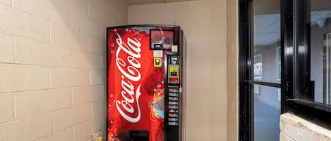 A Coca-Cola vending machine in a corner with a tiled floor and a window with a black frame.
