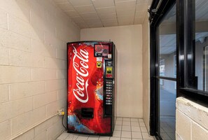 Una máquina expendedora de Coca-Cola en un rincón, con piso de baldosas y una ventana de marco negro.