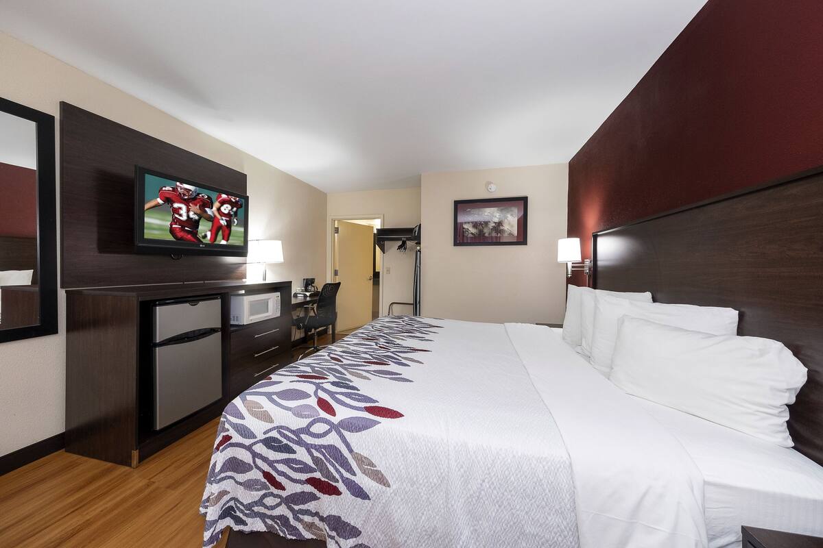 A hotel room with a bed, a desk with a television, a refrigerator, a chair, and a door leading to another room.