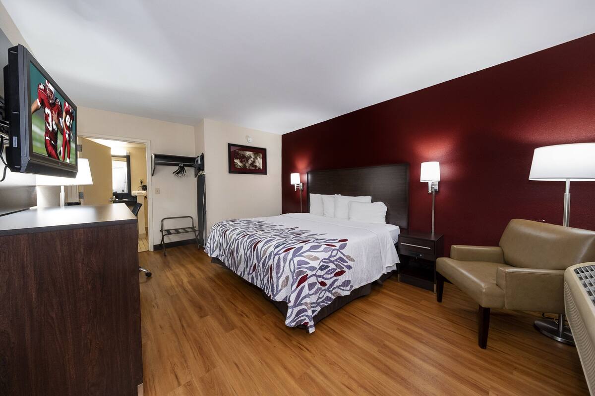 A hotel room with a bed, a desk, a chair, a TV, and a wall-mounted lamp.