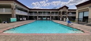 Zona de piscina con una piscina azul cristalina, rodeada por un edificio con múltiples balcones y un paseo techado.
