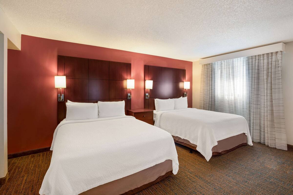 A hotel room with two beds, a large window with curtains, and a red accent wall.
