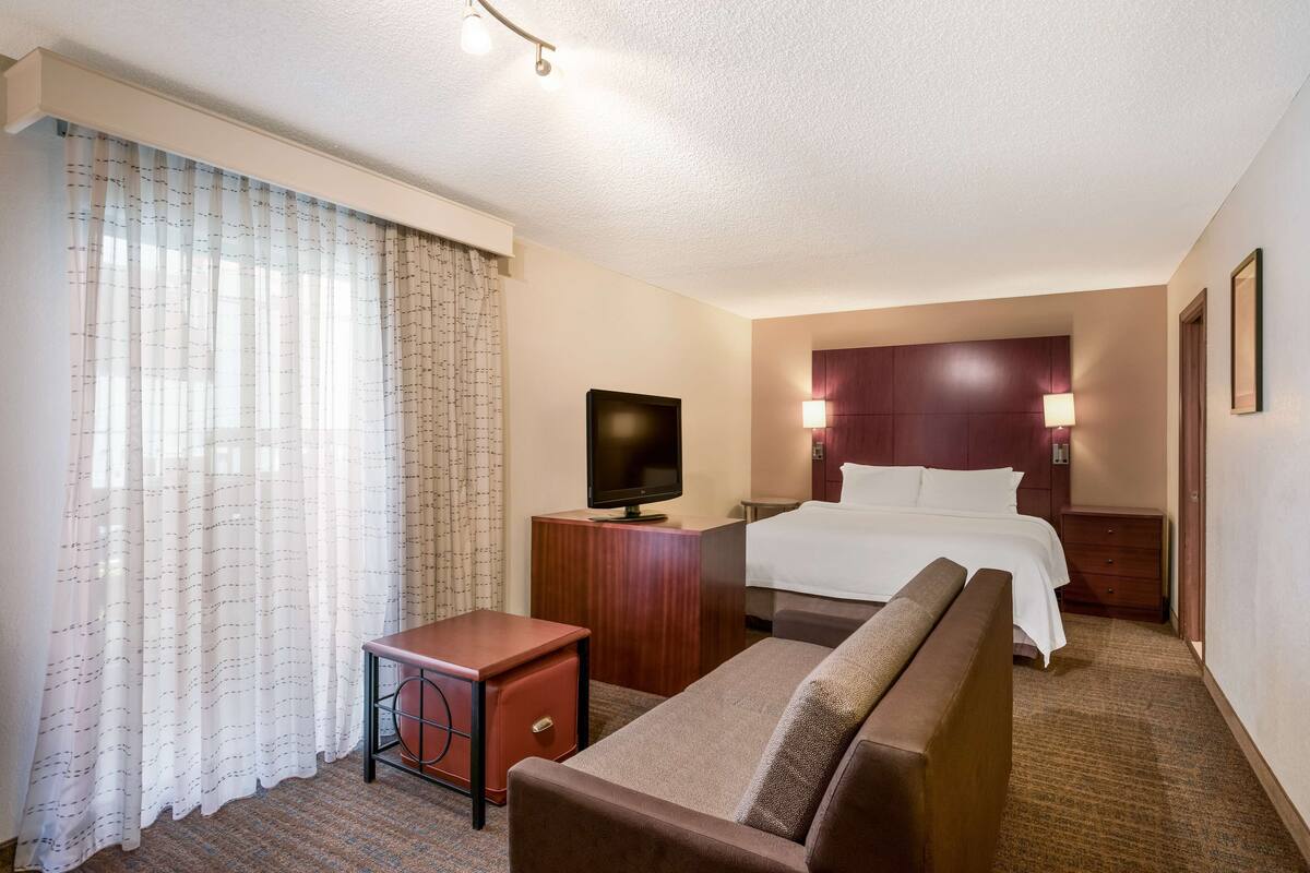 A hotel room with a bed, a TV on a wooden stand, a desk, a chair, and a sofa.