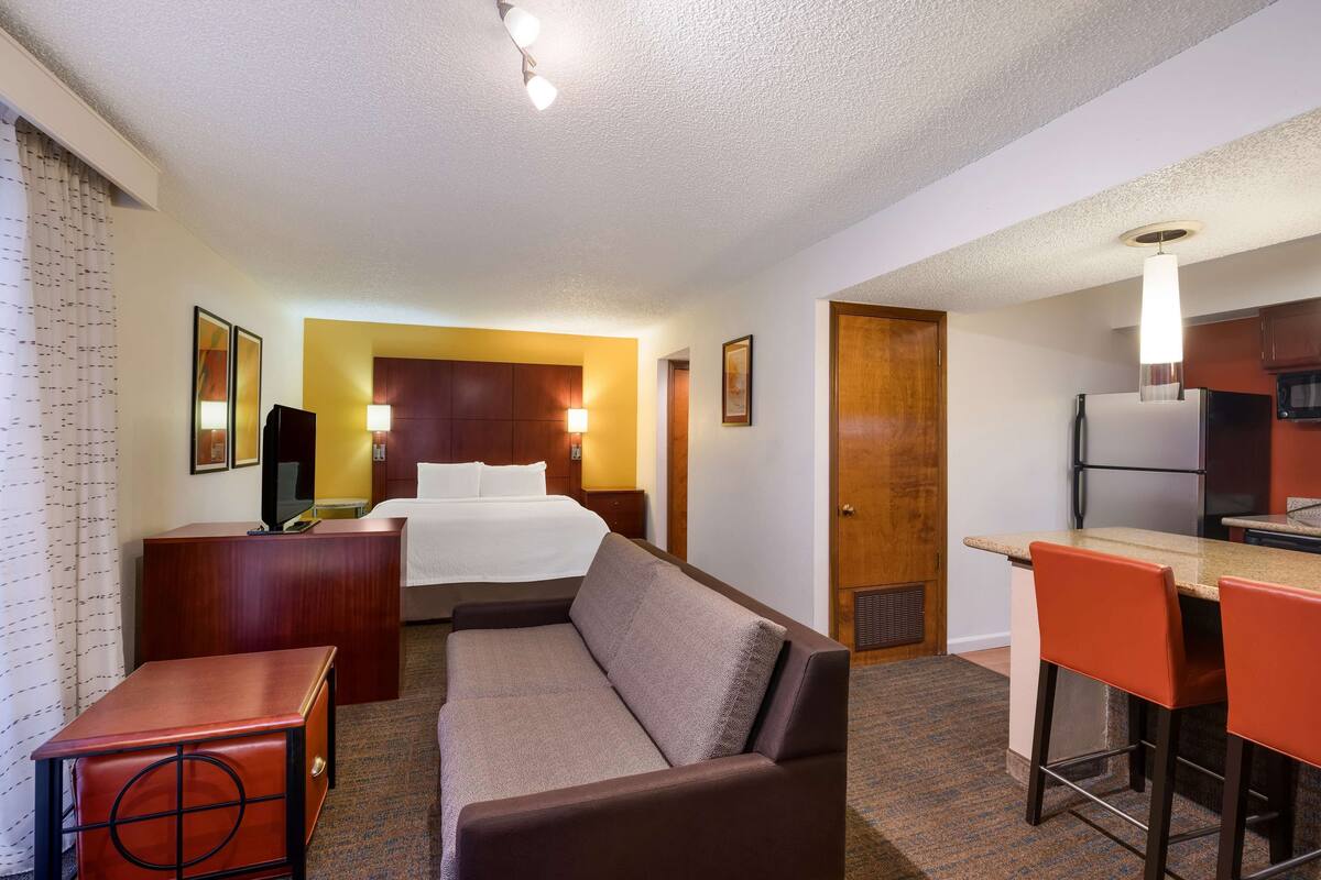 A hotel room with a bed, a sofa, a desk, a kitchenette, and a bathroom.