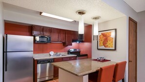 A compact kitchen with a refrigerator, microwave, stove, and dishwasher. There is a small island with two bar stools. The kitchen has wooden cabinets and a red accent wall. - SenS Suites Livermore, SureStay Collection by Best Western (Livermore)