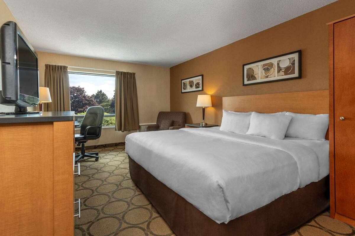 A hotel room with a large bed, a desk with a TV, a chair, two lamps, and a window with curtains.