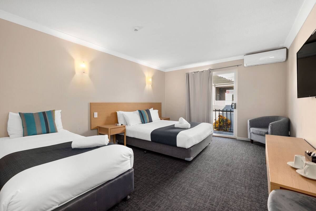Une chambre d’hôtel avec deux lits, un bureau et une chaise. Il y a une fenêtre avec des rideaux, un climatiseur fixé au mur et une porte donnant sur un balcon.