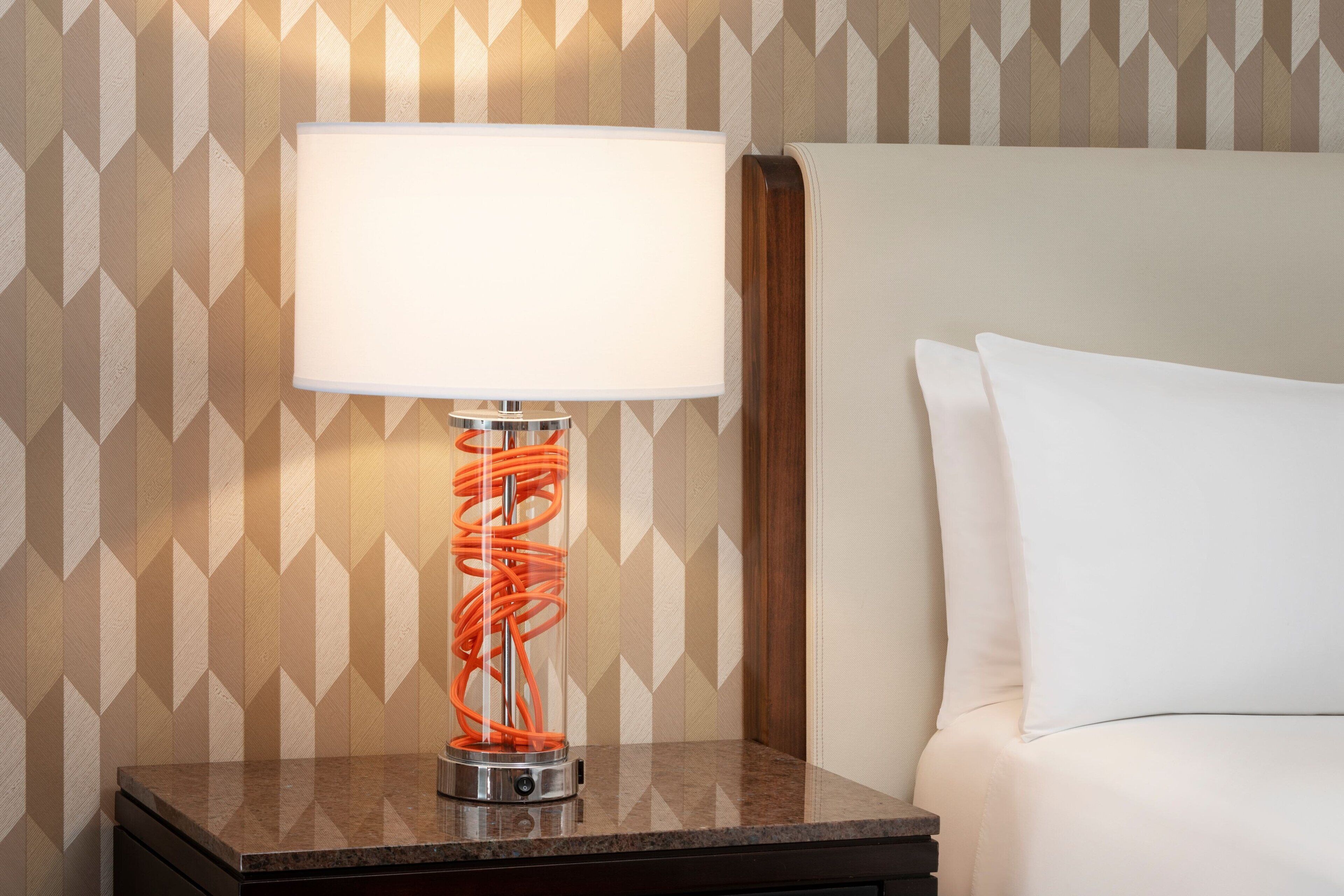 A hotel room with a nightstand, a lamp with a white shade, and a patterned wall.