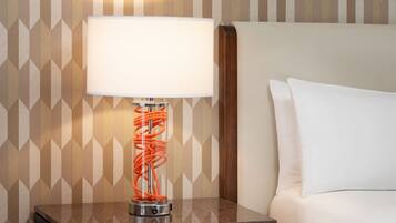 A hotel room with a nightstand, a lamp with a white shade, and a patterned wall.