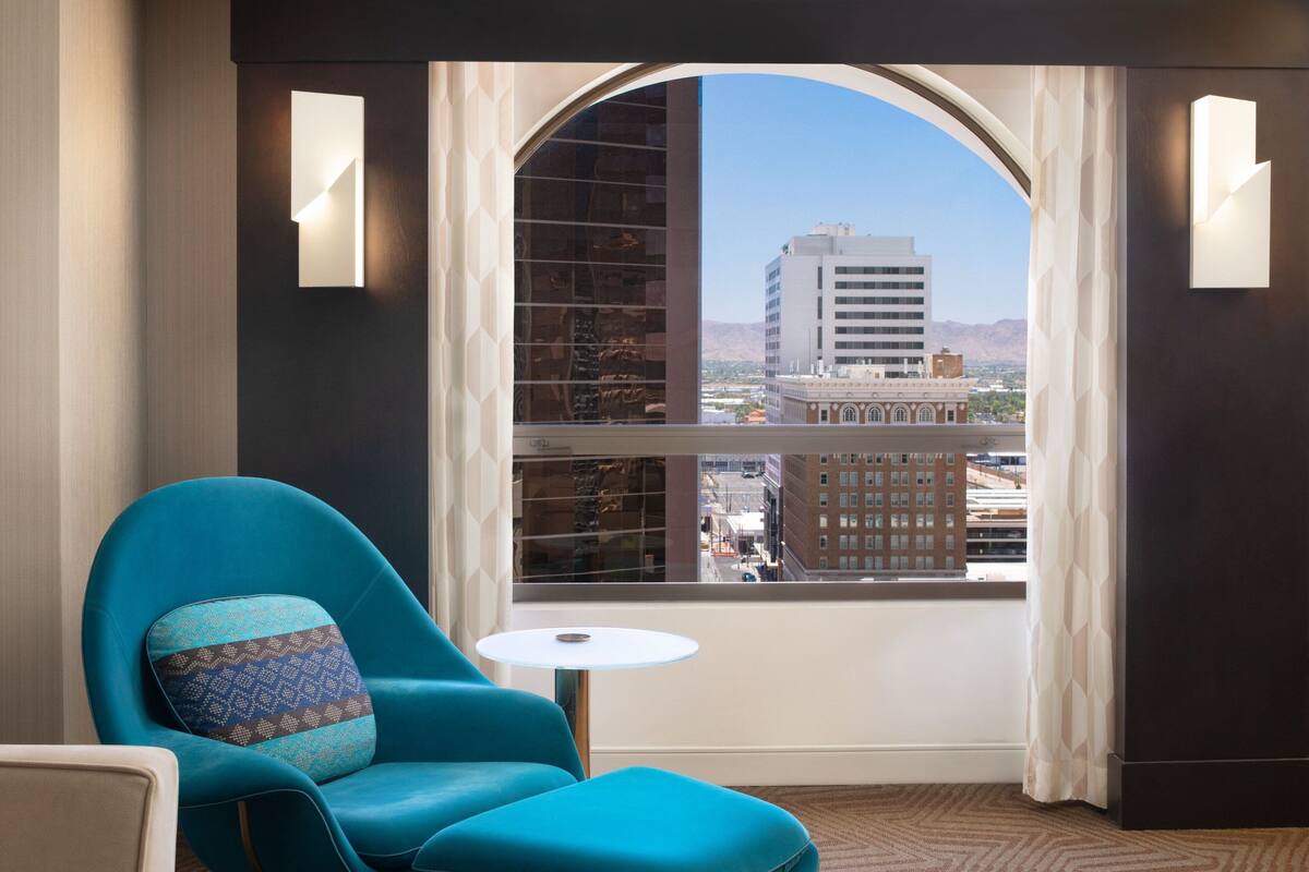 Une pièce dotée d’une grande fenêtre en arc de cercle, d’un fauteuil turquoise agrémenté d’un coussin à motifs, d’une petite table blanche et offrant une vue sur un paysage urbain avec des bâtiments et des montagnes.