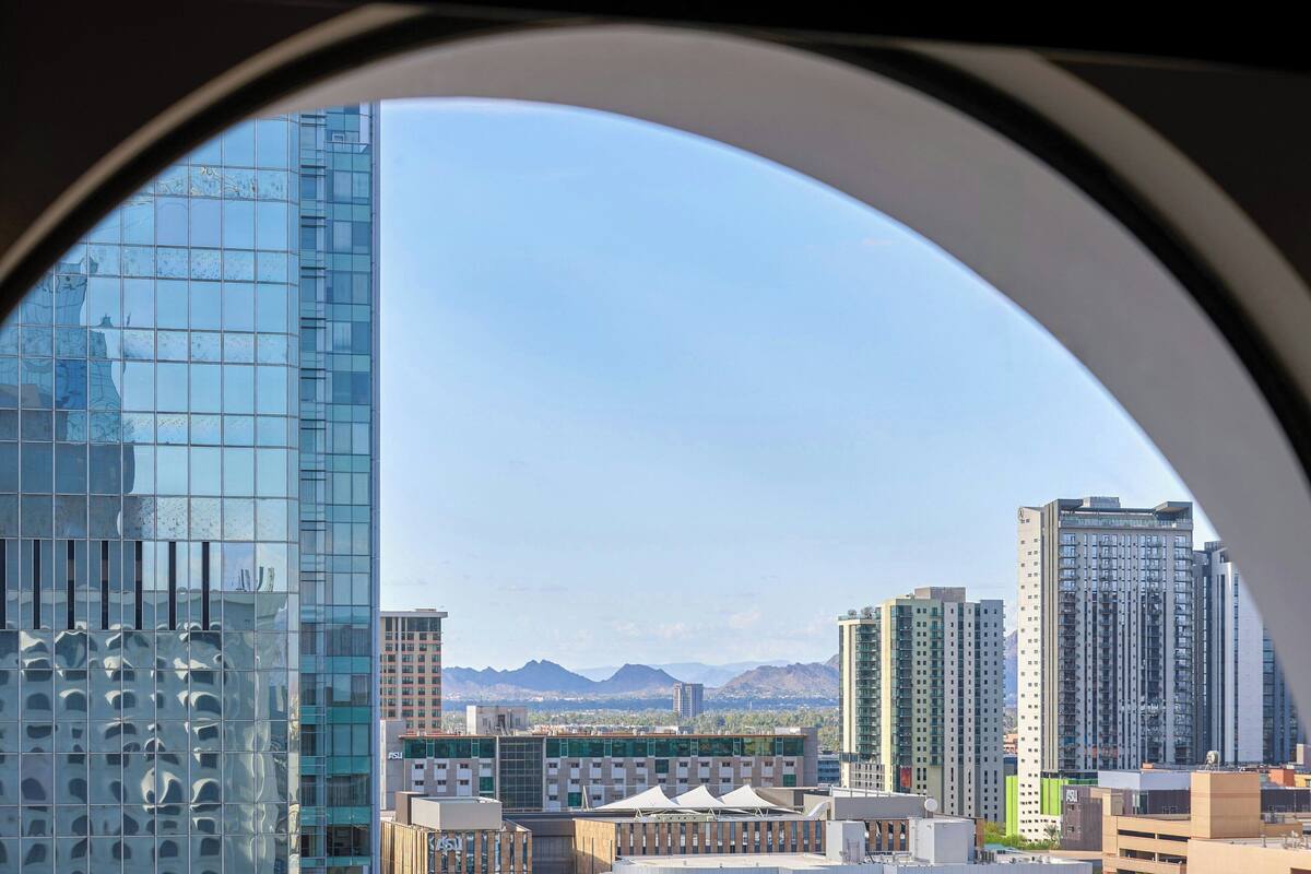 A view through a window of a cityscape with modern buildings, a clear sky, and distant mountains.