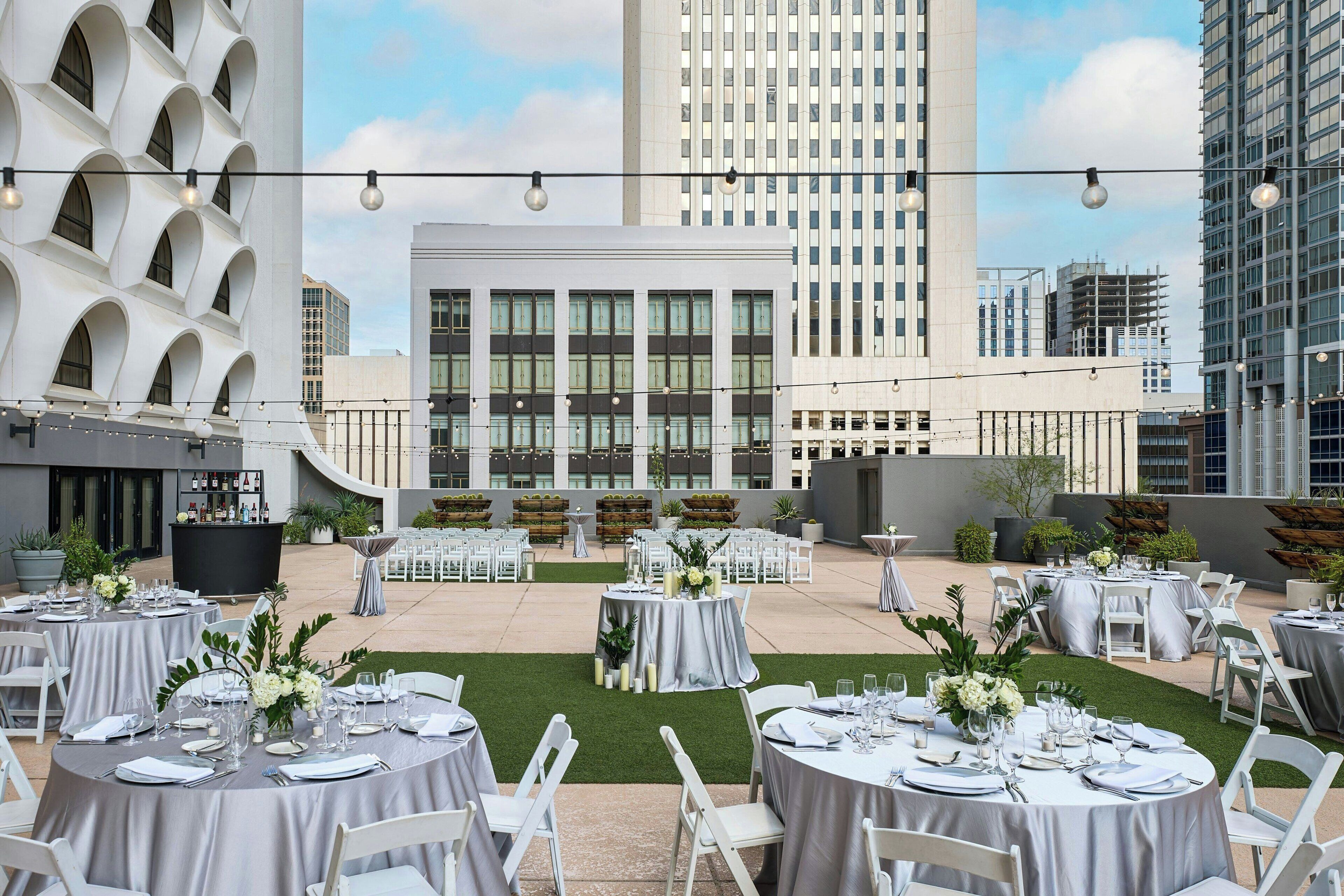 An outdoor dining area with round tables set for a formal event, surrounded by modern buildings and a clear sky.