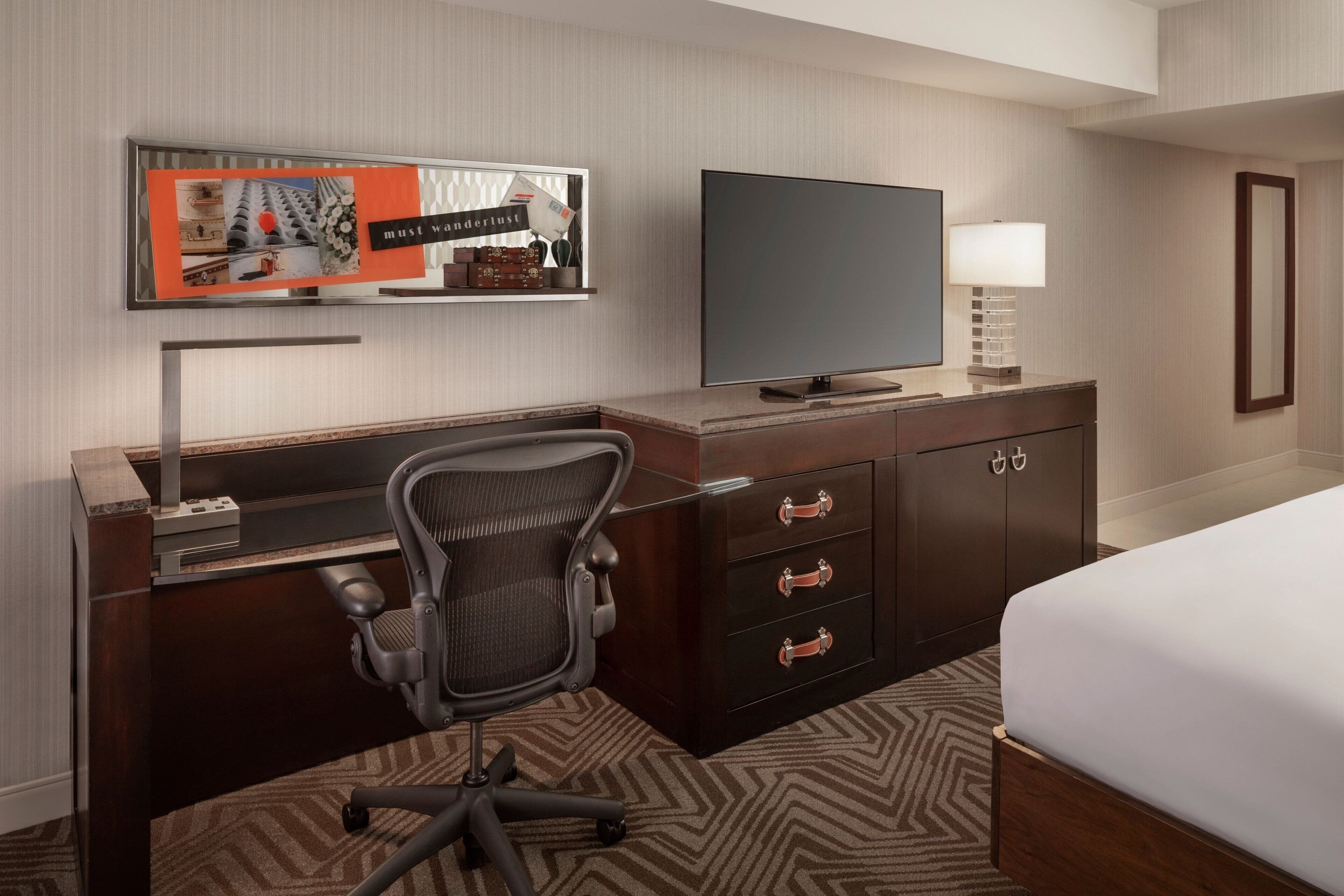 A hotel room with a bed, a desk with a computer, a chair, a TV, and a wall-mounted shelf with a picture frame and a sign.