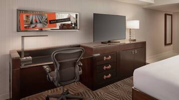 A hotel room with a bed, a desk with a computer, a chair, a TV, and a wall-mounted shelf with a picture frame and a sign.