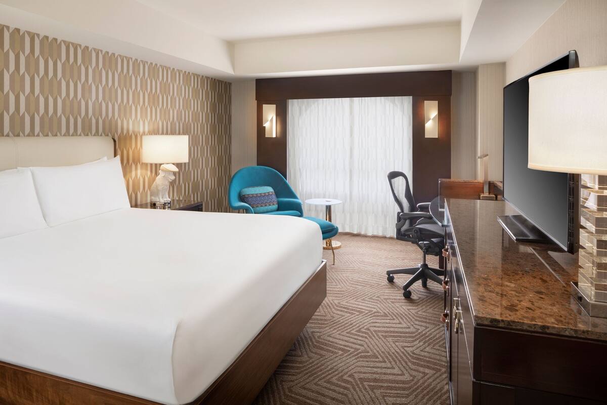 Une chambre d’hôtel avec un grand lit, un bureau doté d’un téléviseur à écran plat, un fauteuil bleu et une petite table ronde.