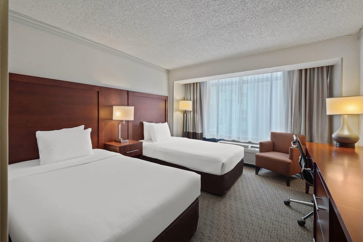 A hotel room with two beds, a desk, a chair, and a large window with curtains.