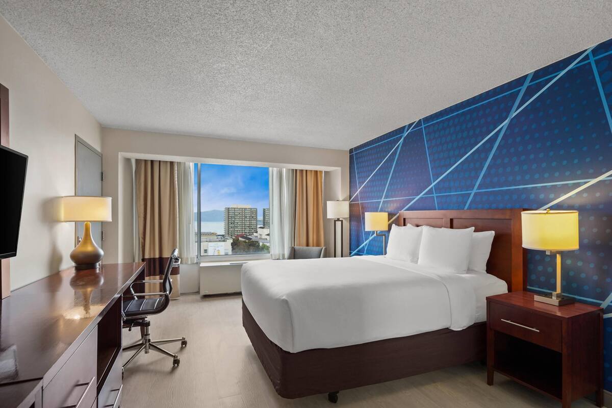 A hotel room with a large bed, a desk with a chair, a TV, and a view of the city.