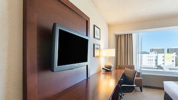 A hotel room with a flat-screen TV mounted on the wall, a desk with a lamp, a chair, and a bed.