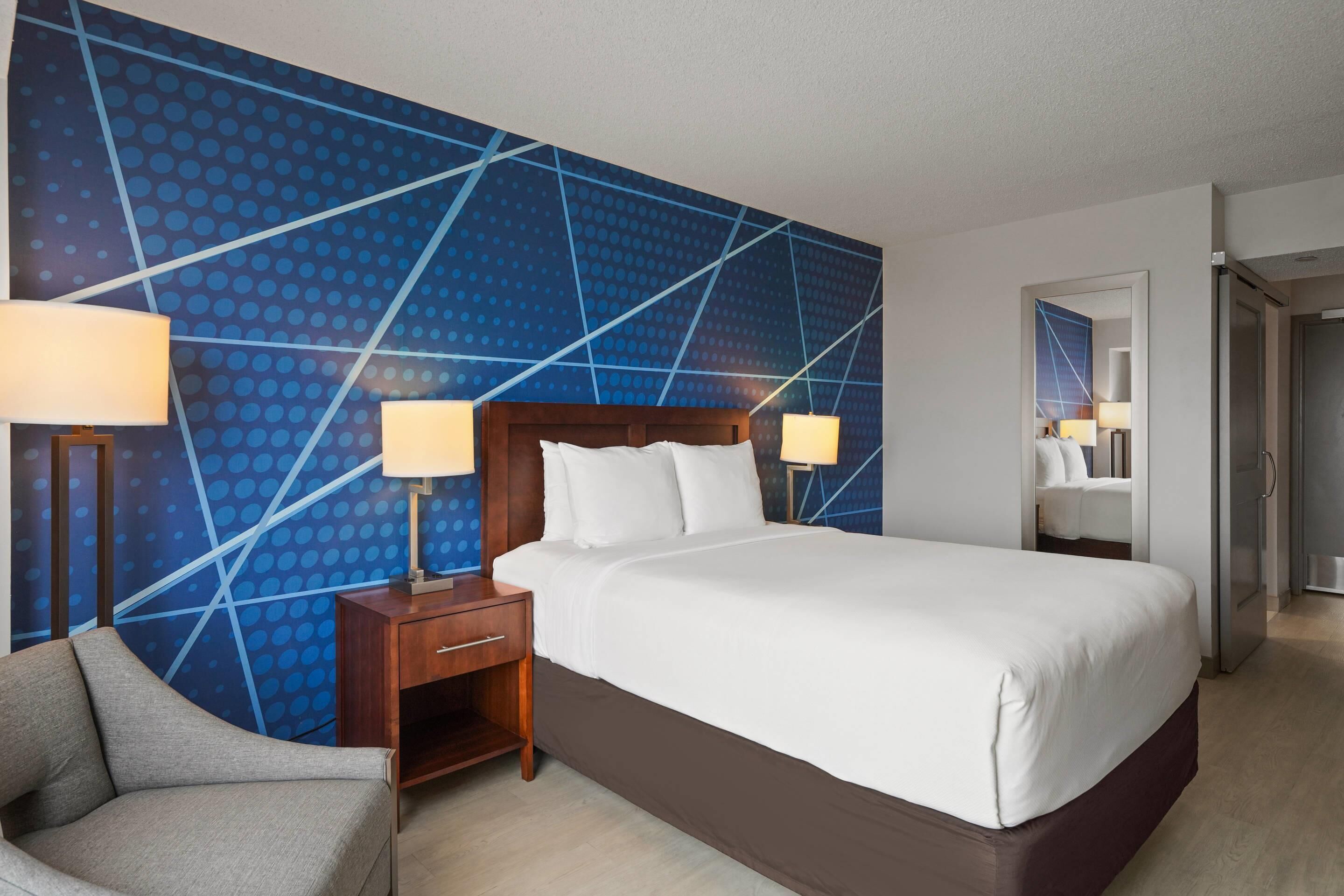 A hotel room with a large bed, two bedside tables with lamps, a chair, and a blue patterned wall.