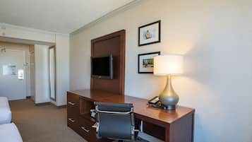 A hotel room with a wooden desk, a chair, a TV mounted on the wall, a lamp, and a bed.
