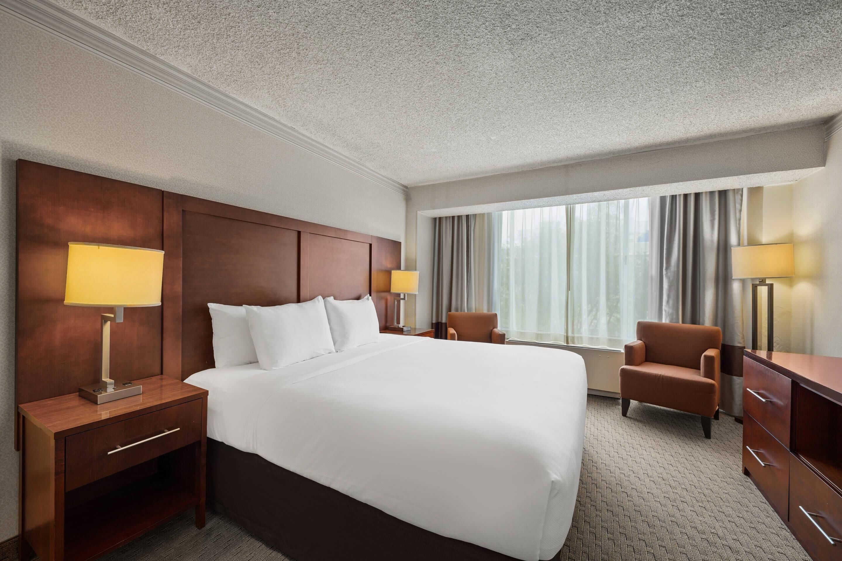 A hotel room with a large bed, two bedside tables with lamps, a chair, a dresser, and a large window with curtains.