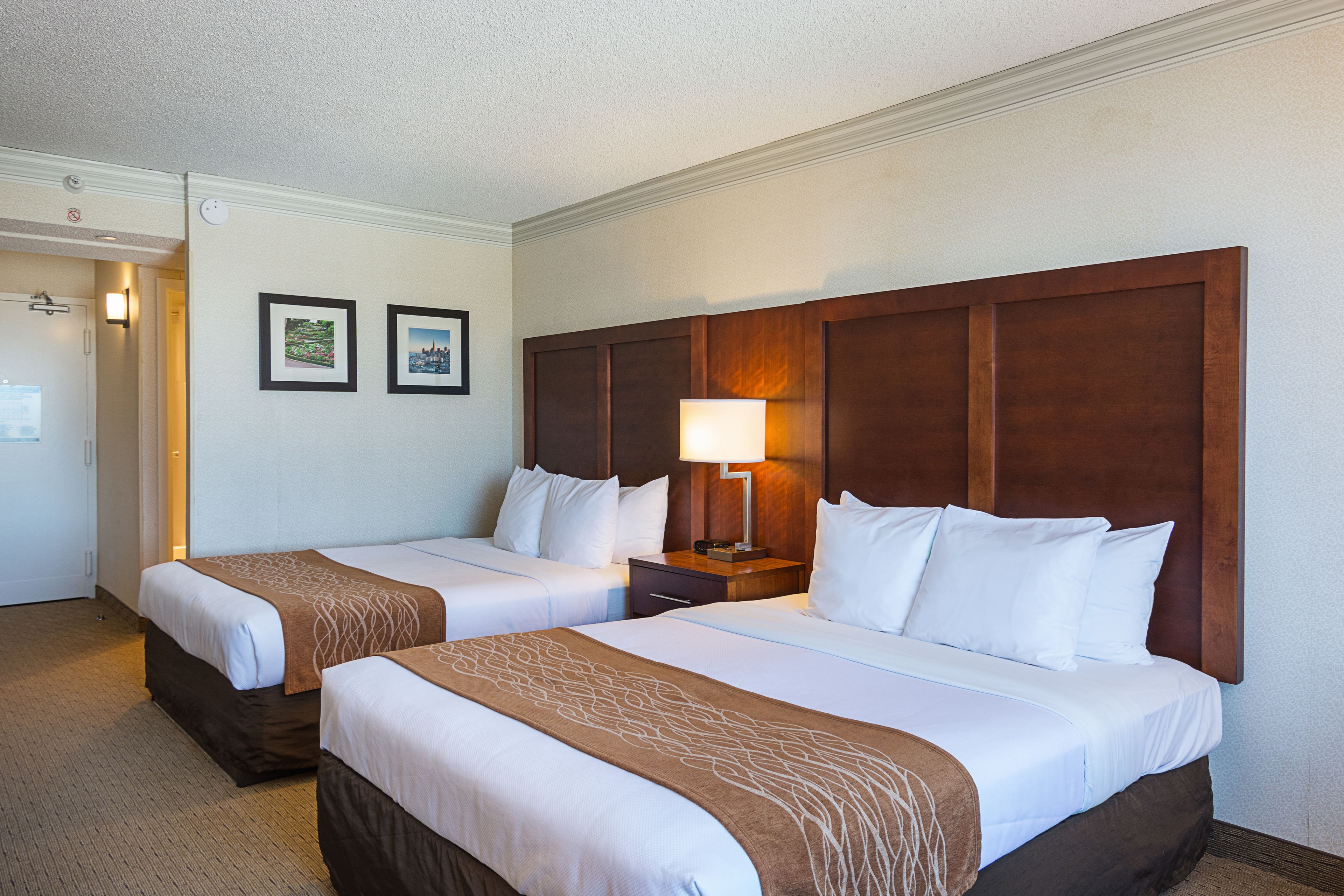 A hotel room with two beds, a wooden headboard, a nightstand with a lamp, and two framed pictures on the wall.