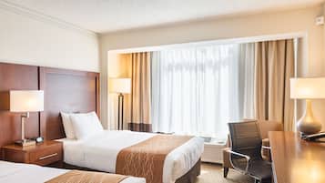 A hotel room with two beds, a desk, and a chair. There are lamps on either side of the beds and a window with curtains.