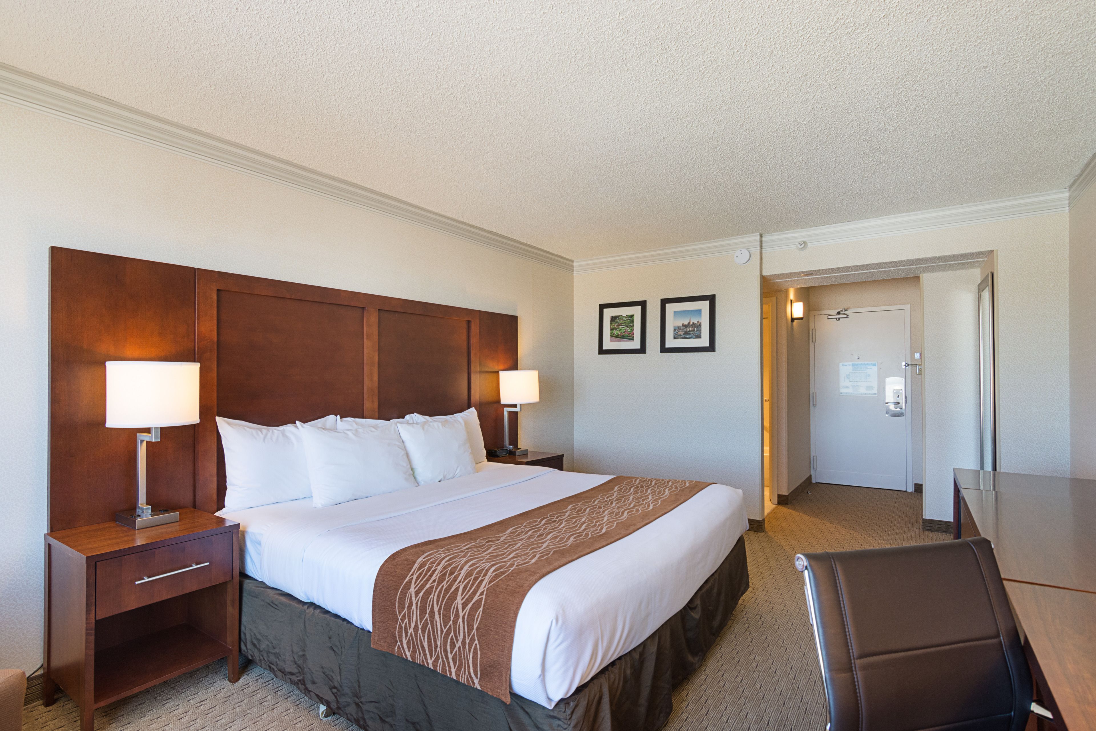 Une chambre d’hôtel avec un grand lit, deux lampes de chevet, une tête de lit en bois, un plaid marron, un bureau avec une chaise et une porte donnant sur une salle de bain.