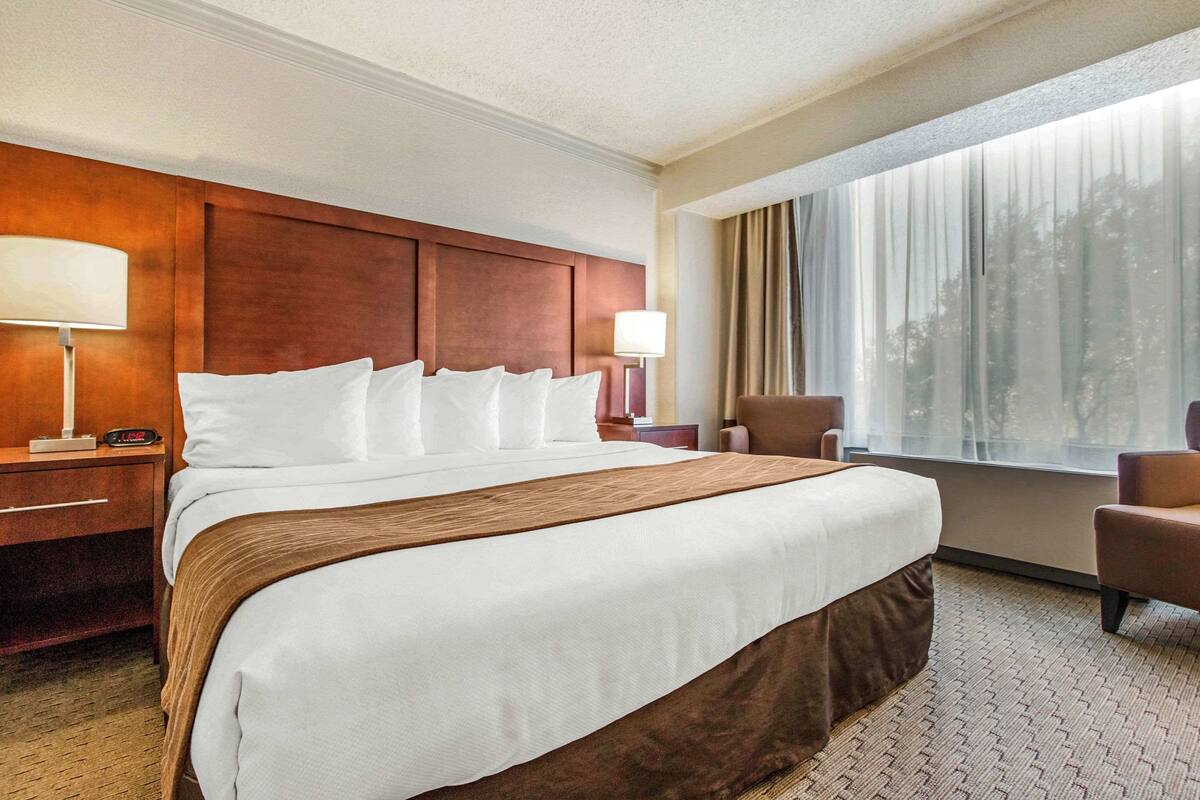 A hotel room with a large bed, a wooden headboard, a nightstand with a lamp, a chair, and a window with curtains.