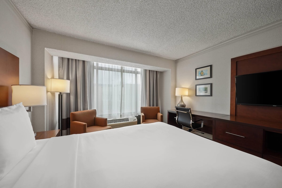 A hotel room with a large bed, two armchairs, a desk with a lamp, a TV, and a window with curtains.