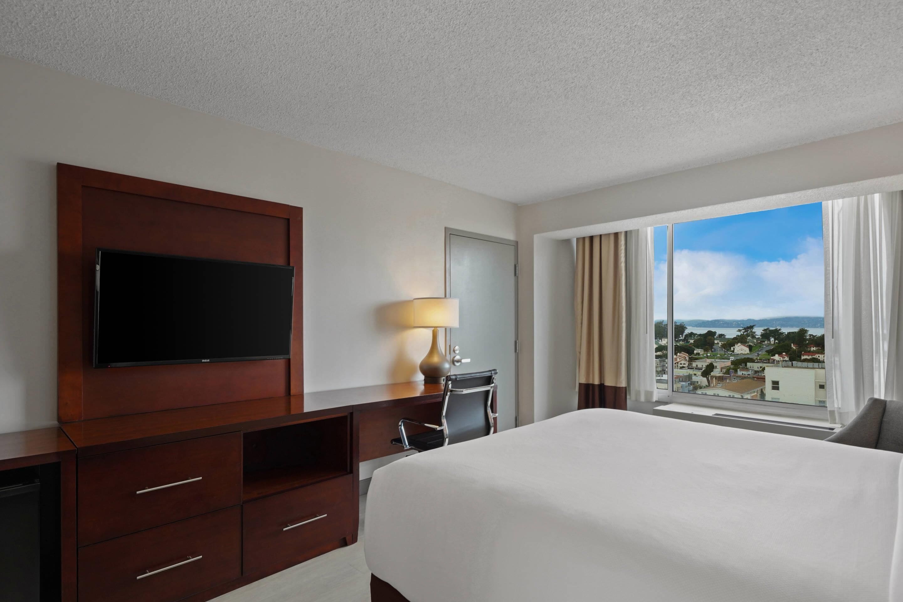 A hotel room with a large bed, a TV on a wooden cabinet, a desk with a lamp, and a window with a city view.